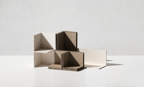A minimalist composition featuring interlocking geometric shapes made from cardboard and white material, positioned on a flat surface with a plain background.