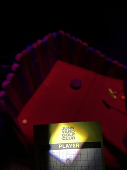 A dimly lit setting with a mini golf course featuring a red surface and a decorative enclosure around it. In the foreground, there is a scorecard with 'Junk Yard Golf Club' visible, and several names with scores written beside them. A small shadow partially obscures the card.