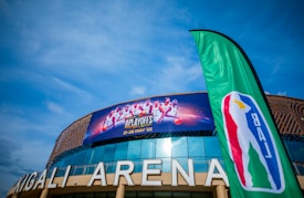Kigali Arena with a large banner advertising a basketball event featuring several players, accompanied by a green flag displaying a basketball-themed logo. The sky is clear and blue, enhancing the vibrant setting.