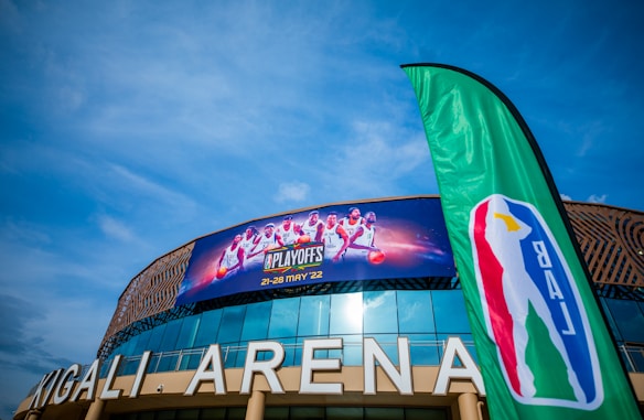 Kigali Arena with a large banner advertising a basketball event featuring several players, accompanied by a green flag displaying a basketball-themed logo. The sky is clear and blue, enhancing the vibrant setting.
