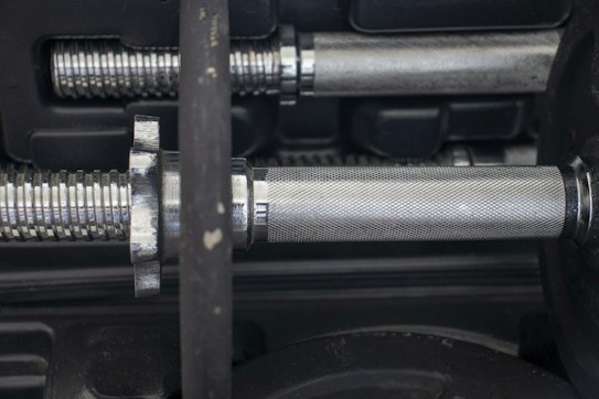 A close-up view of a metal dumbbell handle with a textured grip, surrounded by part of the weight plates. The shiny, metallic surface reflects light, revealing the intricate threading and structure of the dumbbell.