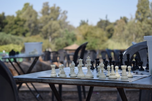 A chess board sits on a metal outdoor table with chess pieces set up for a game. The background is blurred, featuring lush green trees and additional empty tables and chairs, suggesting an outdoor park or garden setting.