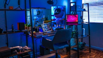 A stylish home office or gaming setup with a futuristic vibe. The room is illuminated with a blue hue, coming from LED lights. On the desk, there's a laptop displaying various apps, surrounded by books and a coffee mug. A small astronaut figurine adds a playful touch, standing next to the laptop. Shelves with plants, accessories, and tech gadgets surround the desk. Blinds partially cover a window on the right, reflecting the soft blue light.