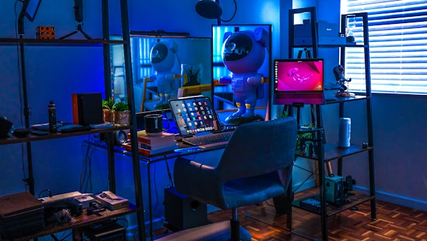A stylish home office or gaming setup with a futuristic vibe. The room is illuminated with a blue hue, coming from LED lights. On the desk, there's a laptop displaying various apps, surrounded by books and a coffee mug. A small astronaut figurine adds a playful touch, standing next to the laptop. Shelves with plants, accessories, and tech gadgets surround the desk. Blinds partially cover a window on the right, reflecting the soft blue light.