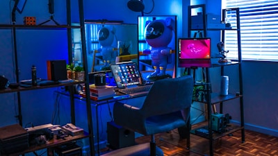 A stylish home office or gaming setup with a futuristic vibe. The room is illuminated with a blue hue, coming from LED lights. On the desk, there's a laptop displaying various apps, surrounded by books and a coffee mug. A small astronaut figurine adds a playful touch, standing next to the laptop. Shelves with plants, accessories, and tech gadgets surround the desk. Blinds partially cover a window on the right, reflecting the soft blue light.