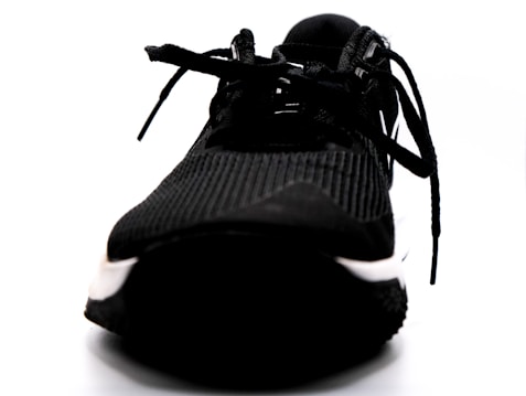 A close-up view of a black athletic shoe with a textured pattern and prominently featured laces. The shoe appears to be placed on a white background, highlighting its dark color and details.
