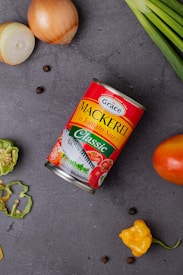 A can of Grace mackerel in tomato sauce lies on a dark gray surface. Surrounding the can are various fresh ingredients including a halved onion, whole onion, green onions, a tomato, a sliced green bell pepper, and scattered black peppercorns. A bright yellow scotch bonnet pepper adds a pop of color.
