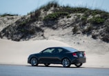 A black car is parked on a road next to sandy dunes with sparse vegetation. The car is positioned to the right side, with grassy and bushy areas visible on the dunes in the background under a clear sky.