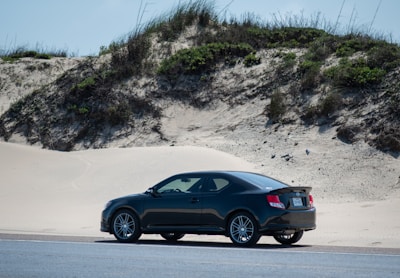 A black car is parked on a road next to sandy dunes with sparse vegetation. The car is positioned to the right side, with grassy and bushy areas visible on the dunes in the background under a clear sky.