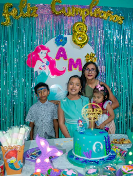 A birthday celebration scene featuring a colorful backdrop with teal fringe curtains, golden balloon letters spelling 'Feliz Cumpleaños', and an '8' balloon, indicating the child's age. The backdrop also includes an image of the Little Mermaid and letters spelling 'AMA'. Four people are posing in front of the backdrop: three children, one of whom is wearing a light blue dress, and an adult holding a younger child. The birthday table is decorated with a blue ocean-themed cake adorned with an '8' and decorated with seaweed, a mermaid topper, and other underwater-themed elements. Additional party items like decorated muffins and a cup showing a Little Mermaid design are visible.