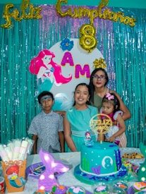 A birthday celebration scene featuring a colorful backdrop with teal fringe curtains, golden balloon letters spelling 'Feliz Cumpleaños', and an '8' balloon, indicating the child's age. The backdrop also includes an image of the Little Mermaid and letters spelling 'AMA'. Four people are posing in front of the backdrop: three children, one of whom is wearing a light blue dress, and an adult holding a younger child. The birthday table is decorated with a blue ocean-themed cake adorned with an '8' and decorated with seaweed, a mermaid topper, and other underwater-themed elements. Additional party items like decorated muffins and a cup showing a Little Mermaid design are visible.