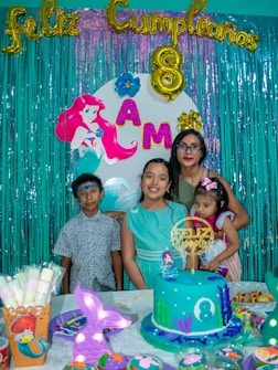 A birthday celebration scene featuring a colorful backdrop with teal fringe curtains, golden balloon letters spelling 'Feliz Cumplea&ntilde;os', and an '8' balloon, indicating the child's age. The backdrop also includes an image of the Little Mermaid and letters spelling 'AMA'. Four people are posing in front of the backdrop: three children, one of whom is wearing a light blue dress, and an adult holding a younger child. The birthday table is decorated with a blue ocean-themed cake adorned with an '8' and decorated with seaweed, a mermaid topper, and other underwater-themed elements. Additional party items like decorated muffins and a cup showing a Little Mermaid design are visible.