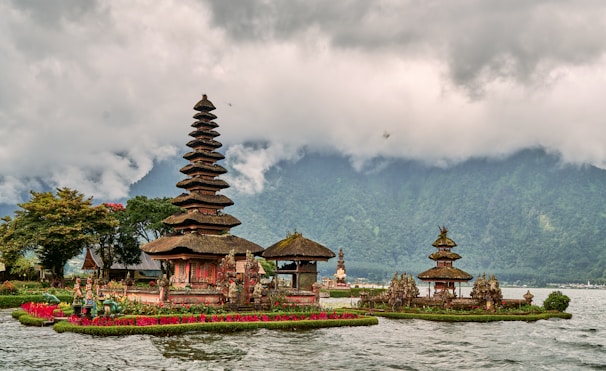 A traditional Balinese temple with tiered towers is situated on the edge of a lake, surrounded by lush greenery and vibrant red flowers. The scene is set against a backdrop of mist-covered mountains and a cloudy sky.