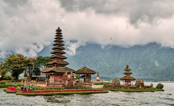 A traditional Balinese temple with tiered towers is situated on the edge of a lake, surrounded by lush greenery and vibrant red flowers. The scene is set against a backdrop of mist-covered mountains and a cloudy sky.