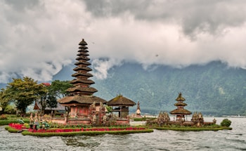 A traditional Balinese temple with tiered towers is situated on the edge of a lake, surrounded by lush greenery and vibrant red flowers. The scene is set against a backdrop of mist-covered mountains and a cloudy sky.