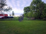 A large satellite dish stands on grass in a rural setting. To the left, there's a red-roofed shed or barn, while lush green trees and a wire fence are visible in the background. The sky is overcast with soft clouds, suggesting a calm day.