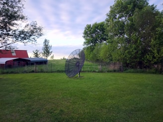 A large satellite dish stands on grass in a rural setting. To the left, there's a red-roofed shed or barn, while lush green trees and a wire fence are visible in the background. The sky is overcast with soft clouds, suggesting a calm day.