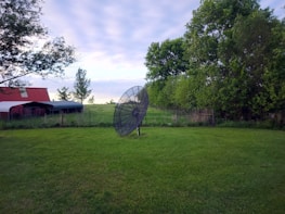 A large satellite dish stands on grass in a rural setting. To the left, there's a red-roofed shed or barn, while lush green trees and a wire fence are visible in the background. The sky is overcast with soft clouds, suggesting a calm day.