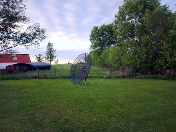 A large satellite dish stands on grass in a rural setting. To the left, there's a red-roofed shed or barn, while lush green trees and a wire fence are visible in the background. The sky is overcast with soft clouds, suggesting a calm day.