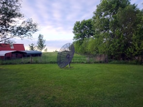 A large satellite dish stands on grass in a rural setting. To the left, there's a red-roofed shed or barn, while lush green trees and a wire fence are visible in the background. The sky is overcast with soft clouds, suggesting a calm day.