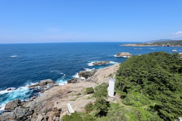 A picturesque coastline with clear blue skies and a vast expanse of ocean stretching to the horizon. Rugged rocks and waves crashing against them create a dynamic shoreline. A white lighthouse is nestled amidst lush green trees on the right side, adding a charming focal point to the natural scenery.