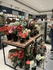 A variety of artificial flowers in vibrant colors are arranged in black pots on a wooden and metal display table within a home décor store. The shelves in the background are filled with pillows and other decorative items. A person in a casual outfit is partially visible on the right side, holding an item and examining merchandise.