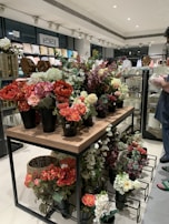A variety of artificial flowers in vibrant colors are arranged in black pots on a wooden and metal display table within a home décor store. The shelves in the background are filled with pillows and other decorative items. A person in a casual outfit is partially visible on the right side, holding an item and examining merchandise.