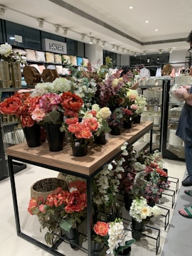 A variety of artificial flowers in vibrant colors are arranged in black pots on a wooden and metal display table within a home décor store. The shelves in the background are filled with pillows and other decorative items. A person in a casual outfit is partially visible on the right side, holding an item and examining merchandise.