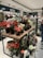 A variety of artificial flowers in vibrant colors are arranged in black pots on a wooden and metal display table within a home décor store. The shelves in the background are filled with pillows and other decorative items. A person in a casual outfit is partially visible on the right side, holding an item and examining merchandise.