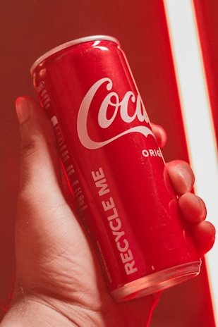 A person's hand holds a red Coca-Cola can with the words 'Recycle Me' printed vertically on the side. The background and lighting are tinted red, emphasizing the color of the can.