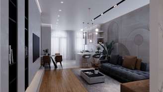 A modern and stylish living room with an open concept design. The space includes a comfortable dark gray sectional sofa with brown cushions, a low rectangular coffee table with a decorative item on top, and a lush green plant beside the sofa. The room has a wooden floor in the foreground, transitioning to a tiled floor towards the back. A dining area with a sleek table and chairs can be seen near large windows adorned with sheer white curtains, allowing natural light to fill the space. Above, modern pendant lights hang from the ceiling, and shelves with decorative items are mounted on the far wall.