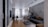 A modern and stylish living room with an open concept design. The space includes a comfortable dark gray sectional sofa with brown cushions, a low rectangular coffee table with a decorative item on top, and a lush green plant beside the sofa. The room has a wooden floor in the foreground, transitioning to a tiled floor towards the back. A dining area with a sleek table and chairs can be seen near large windows adorned with sheer white curtains, allowing natural light to fill the space. Above, modern pendant lights hang from the ceiling, and shelves with decorative items are mounted on the far wall.