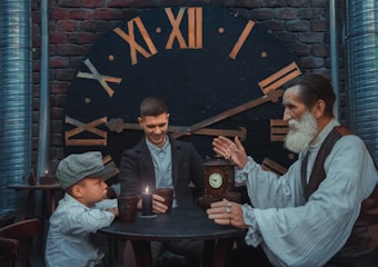 Three people sit around a small, round table with a lit candle atop it, casting soft light on their faces. A large vintage clock face in the background features Roman numerals, set against a brick wall. A young boy in a striped cap, a middle-aged man smiling warmly, and an elderly man with a long beard, dressed in vintage attire, are engaged in what appears to be a storytelling session. The elder is gesturing expressively with his hands.