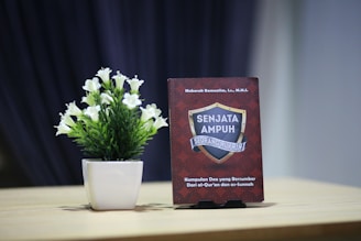 A small potted plant with white flowers is placed on a wooden table next to an upright book. The book has a red cover with a decorative pattern and features a shield emblem with the text 'SENJATA AMPUH SEORANG MUKMIN'. The background is a dark blurred curtain, creating a contrast with the lighter tones of the table and plant.