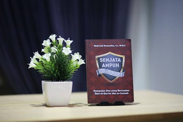 A small potted plant with white flowers is placed on a wooden table next to an upright book. The book has a red cover with a decorative pattern and features a shield emblem with the text 'SENJATA AMPUH SEORANG MUKMIN'. The background is a dark blurred curtain, creating a contrast with the lighter tones of the table and plant.