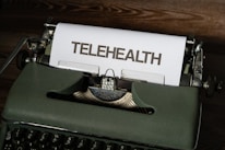 A vintage typewriter with green housing holds a sheet of paper with the word 'TELEHEALTH' typed on it. The scene is set on a wooden surface.