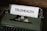 A vintage typewriter with green housing holds a sheet of paper with the word 'TELEHEALTH' typed on it. The scene is set on a wooden surface.
