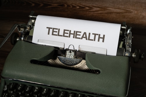 A vintage typewriter with green housing holds a sheet of paper with the word 'TELEHEALTH' typed on it. The scene is set on a wooden surface.