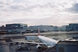 An airplane from Turkish Airlines is parked at an airport gate, surrounded by various ground service vehicles. The terminal building stretches across the background under a partly cloudy sky.