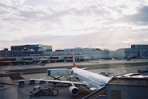 An airplane from Turkish Airlines is parked at an airport gate, surrounded by various ground service vehicles. The terminal building stretches across the background under a partly cloudy sky.