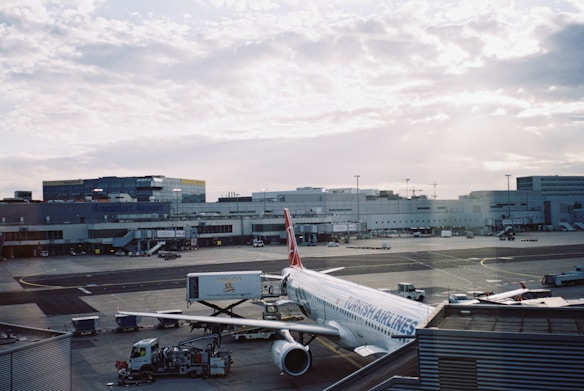 An airplane from Turkish Airlines is parked at an airport gate, surrounded by various ground service vehicles. The terminal building stretches across the background under a partly cloudy sky.