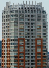 A high-rise building with a modern architectural design featuring a grid of windows and a mix of beige and brown tones. The structure has a distinctive top with vertical protrusions and a rooftop area that may include utility equipment.