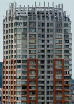 A high-rise building with a modern architectural design featuring a grid of windows and a mix of beige and brown tones. The structure has a distinctive top with vertical protrusions and a rooftop area that may include utility equipment.