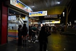 A group of people stands outside a brightly lit crispy chicken shop at night. The storefront features colorful signage in both English and Chinese, indicating a focus on crispy chicken dishes. The setting appears to be on a sidewalk with additional shops visible further down, and the atmosphere is bustling with night activity.