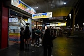 A group of people stands outside a brightly lit crispy chicken shop at night. The storefront features colorful signage in both English and Chinese, indicating a focus on crispy chicken dishes. The setting appears to be on a sidewalk with additional shops visible further down, and the atmosphere is bustling with night activity.