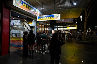 A group of people stands outside a brightly lit crispy chicken shop at night. The storefront features colorful signage in both English and Chinese, indicating a focus on crispy chicken dishes. The setting appears to be on a sidewalk with additional shops visible further down, and the atmosphere is bustling with night activity.