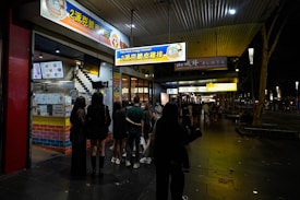 A group of people stands outside a brightly lit crispy chicken shop at night. The storefront features colorful signage in both English and Chinese, indicating a focus on crispy chicken dishes. The setting appears to be on a sidewalk with additional shops visible further down, and the atmosphere is bustling with night activity.