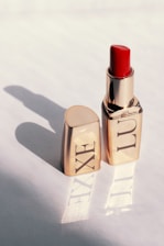 A gold-colored lipstick tube stands upright with the cap placed beside it on a light surface. The lipstick itself is a vibrant red, and the light casts a shadow of the tube and cap on the surface, reflecting the word 'LUXE' from the packaging.