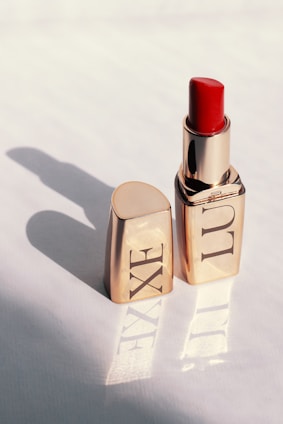A gold-colored lipstick tube stands upright with the cap placed beside it on a light surface. The lipstick itself is a vibrant red, and the light casts a shadow of the tube and cap on the surface, reflecting the word 'LUXE' from the packaging.