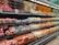A supermarket produce section displays a neatly organized assortment of fruits. The shelves are filled with apples, grapes, pears, oranges, and other fruits, each packed in individual wrapping or foam nets. Pricing labels are visible above the fruits, indicating price per kilogram.